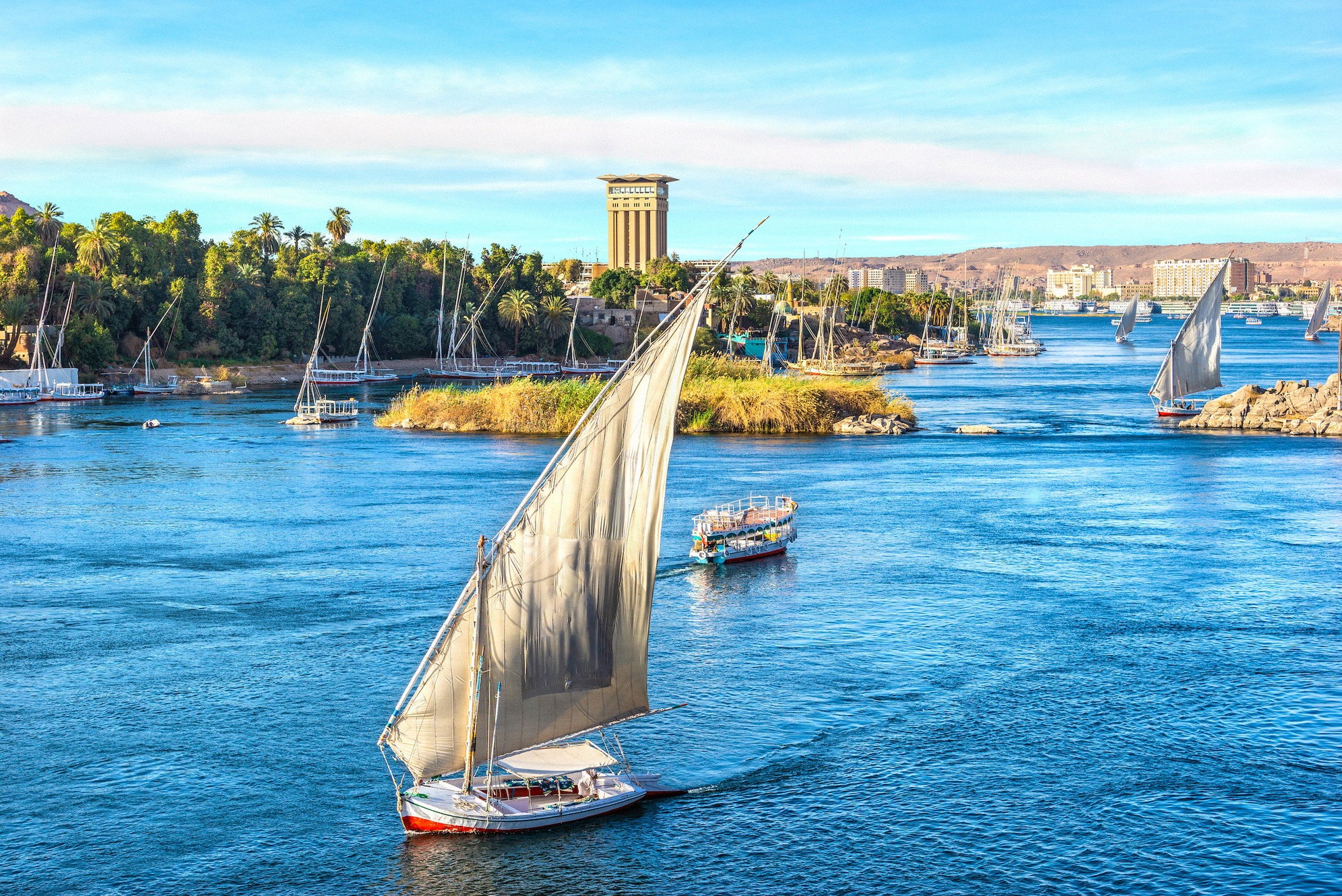 Perinteinen egyptiläinen felucca purjehtii auringonpaisteessa Niilillä Assuanissa, ympärillä muita purjeveneitä, reheviä Niilin saaria ja rannan kaupunkisilhuetti.