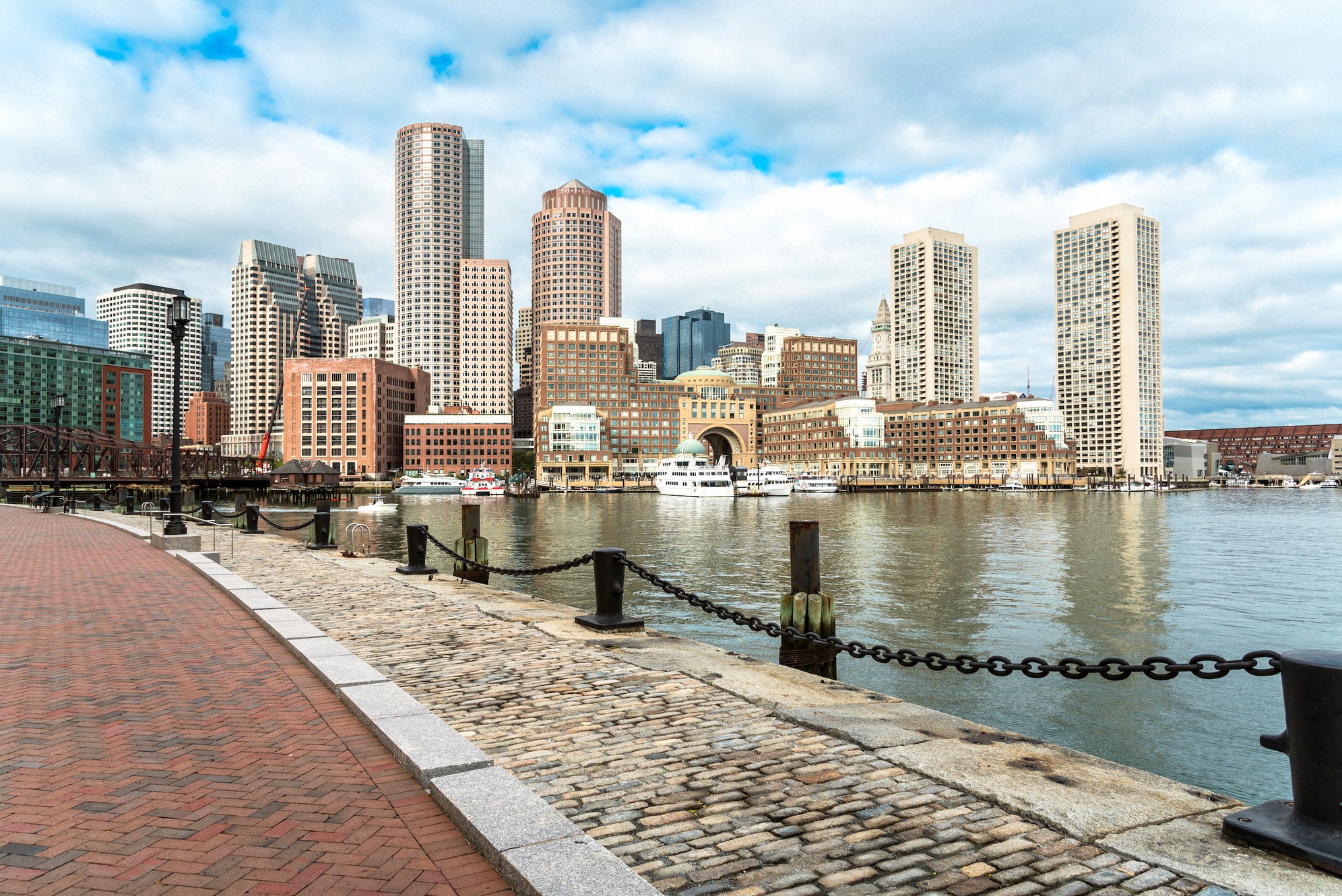Näkymä mukulakivetyltä satamapolulta Bostonin siluettiin, pilvenpiirtäjiä, lauttalaitureita ja tyyntä vettä pilvisenä syysaamuna.