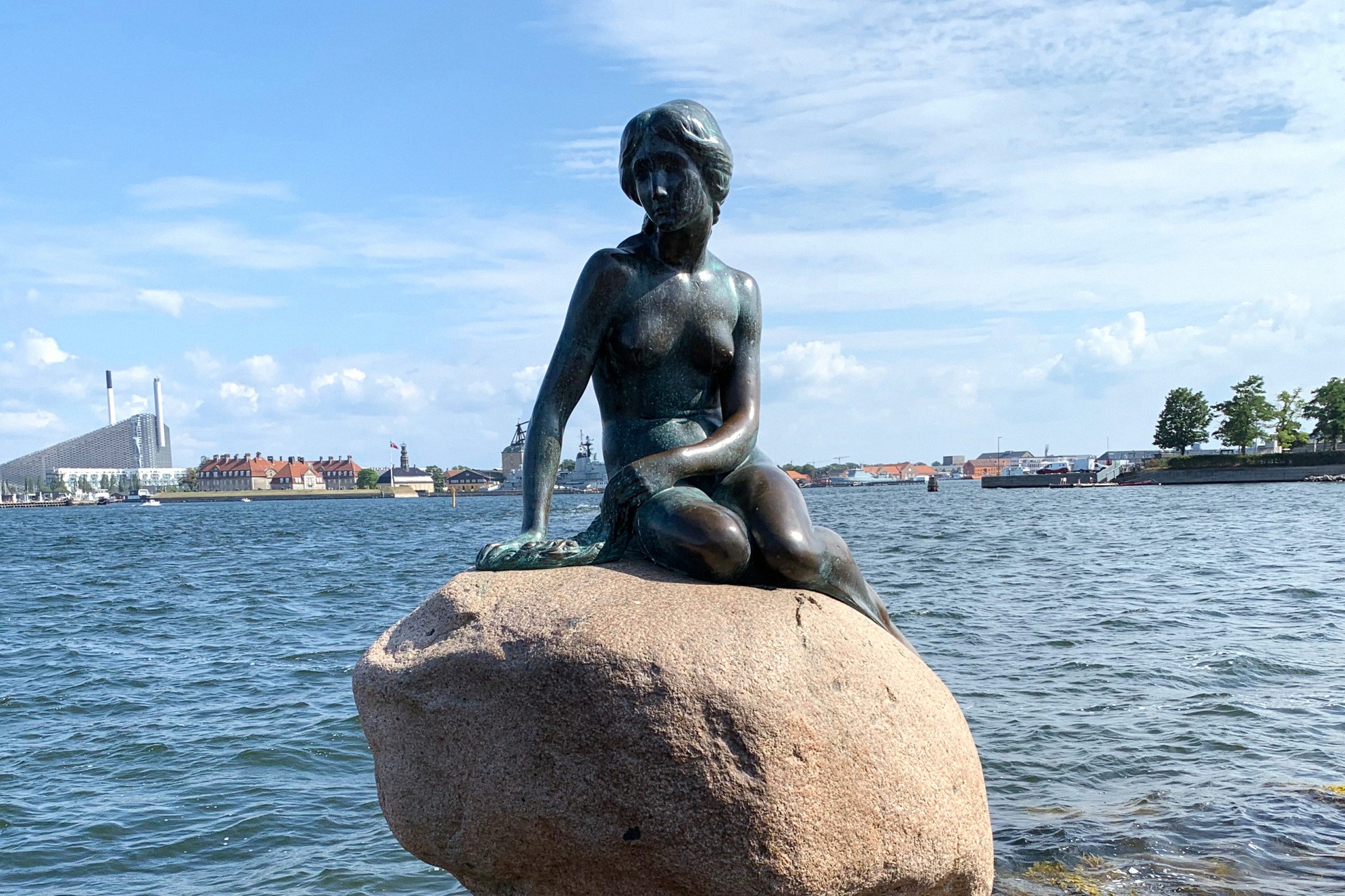 Maailmankuulu Pieni merenneito -pronssipatsas istuu kivellä Kööpenhaminan satamassa, taustalla kaupungin siluetti.
