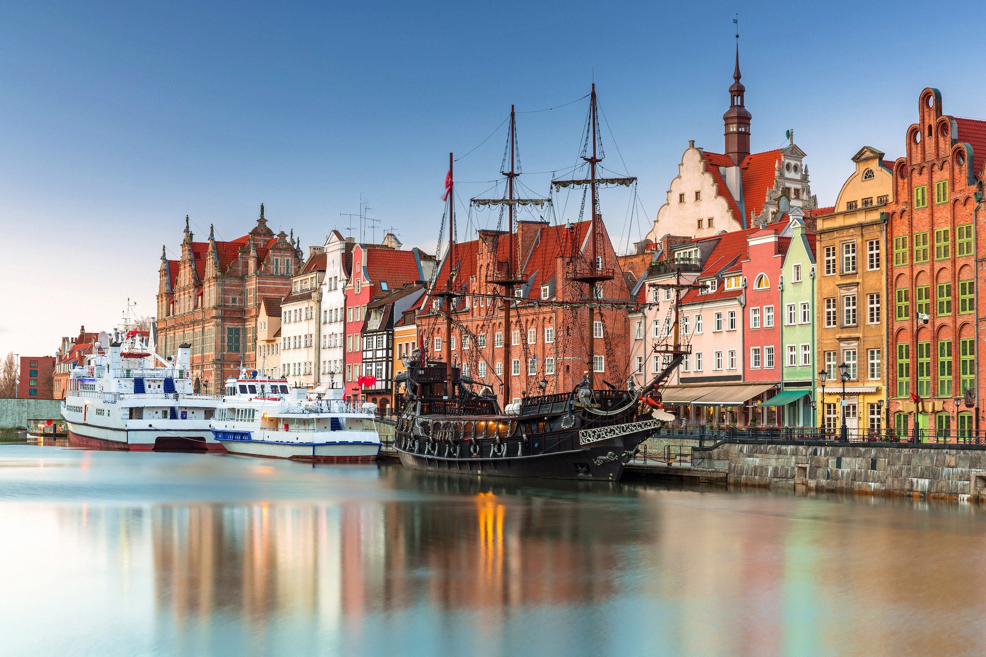 Peilityyni Motława-joki Gdańskin historiallisen vanhankaupungin (Długie Pobrzeże) edessä iltahämärässä, kuvassa merirosvolaivan jäljitelmä ja värikkäitä hansataloja.