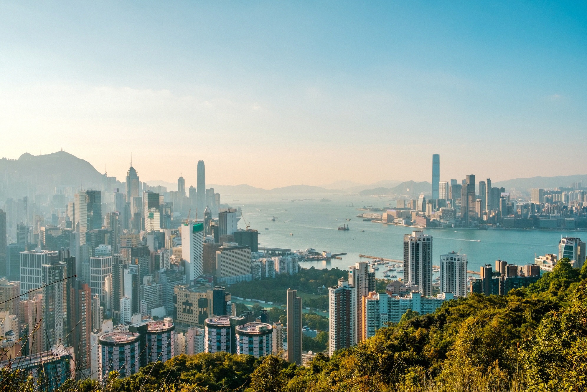 Panoraama Hongkong Islandin ja Kowloonin pilvenpiirtäjistä Victoria Harbourin yli kultaisessa aamuvalossa, tornit ja satama heijastavat kaupungin sykettä.