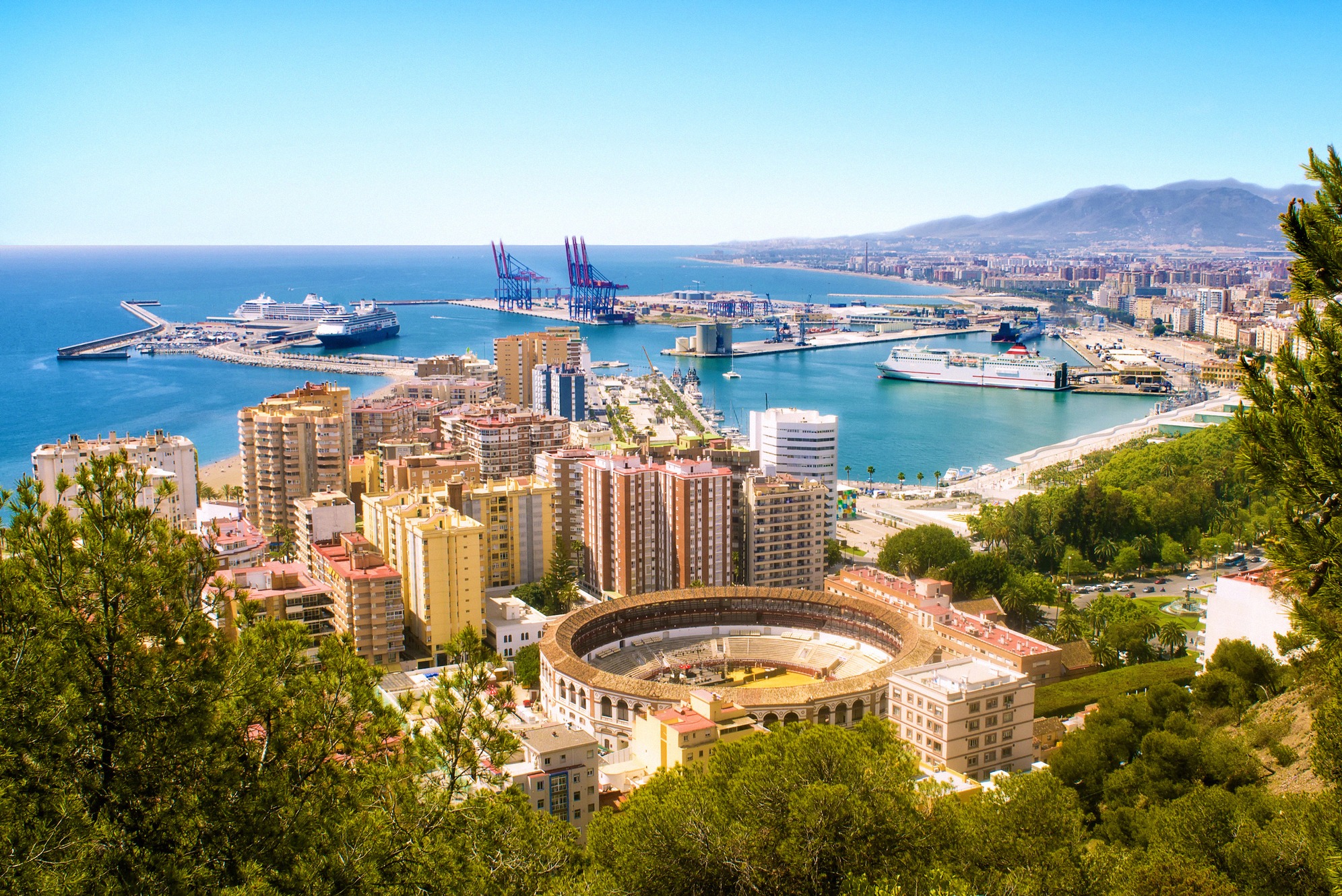 Panoraamanäkymä Gibralfaron rinteiltä Málagaan: etualalla pyöreä härkätaisteluareena La Malagueta, sen takana suuri konttisatama risteilyaluksineen ja sininen Välimeri horisontissa.