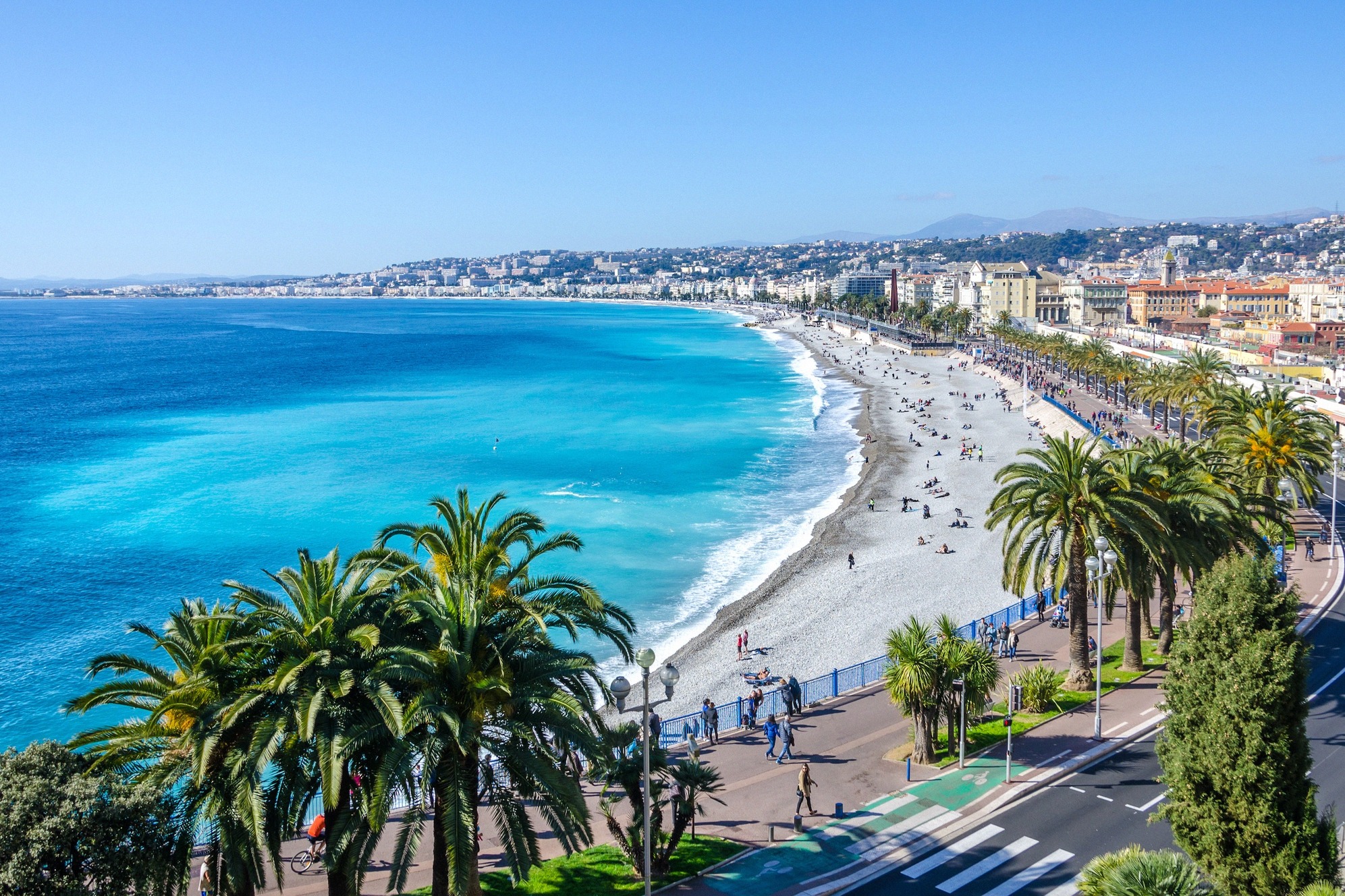 Panoraamakuva Nizzan kuuluisasta Promenade des Anglais'sta: kirkkaansininen Välimeri, kivikkoinen ranta ja palmujen reunustama rantabulevardi täynnä kävelijöitä.
