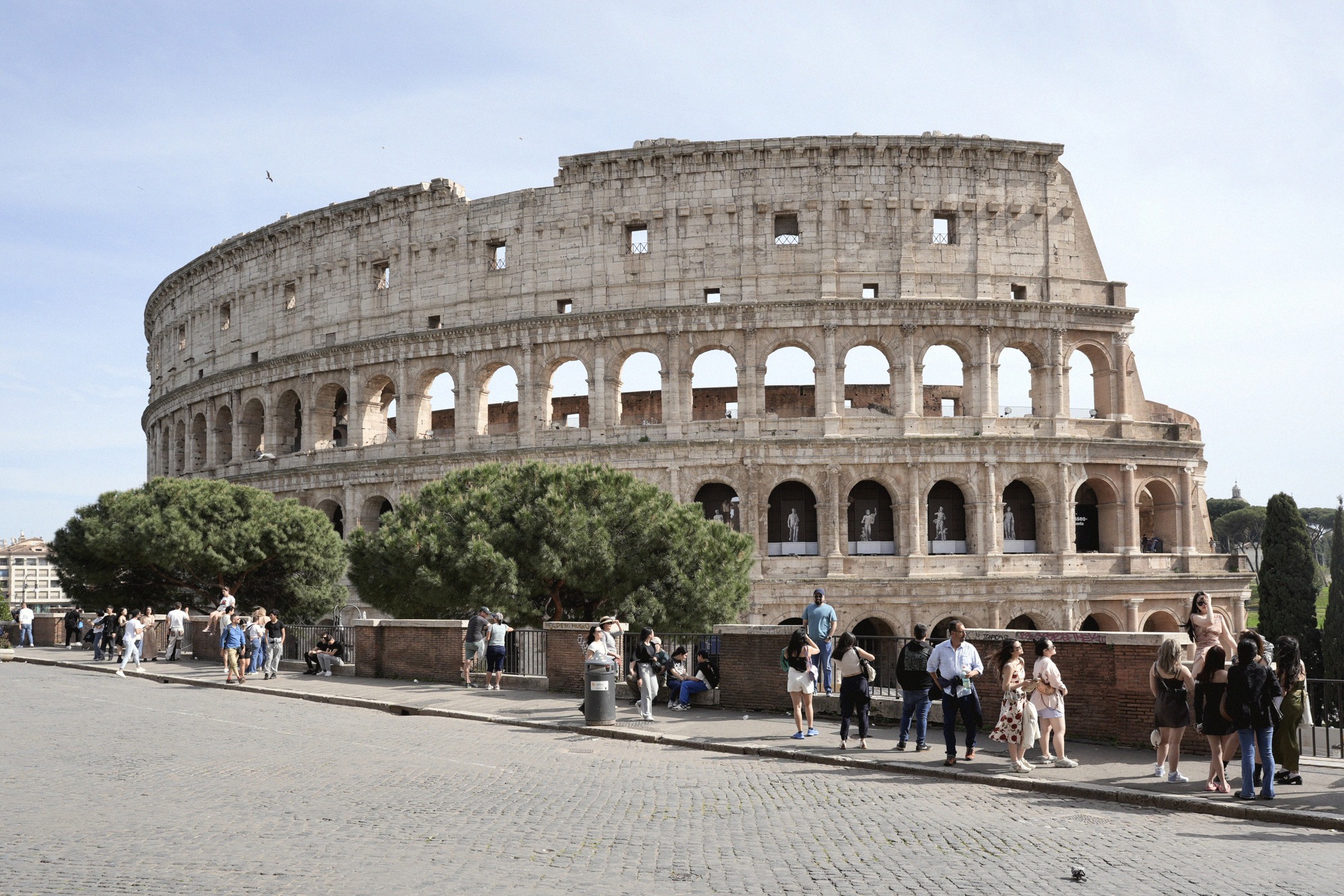 Colosseum Roomassa ulkoa kuvattuna aurinkoisena päivänä. Kivetyllä aukiolla kulkee runsaasti matkailijoita, ja antiikin amfiteatterin kaarineen ja holvikäytävineen hallitsee kuvaa.