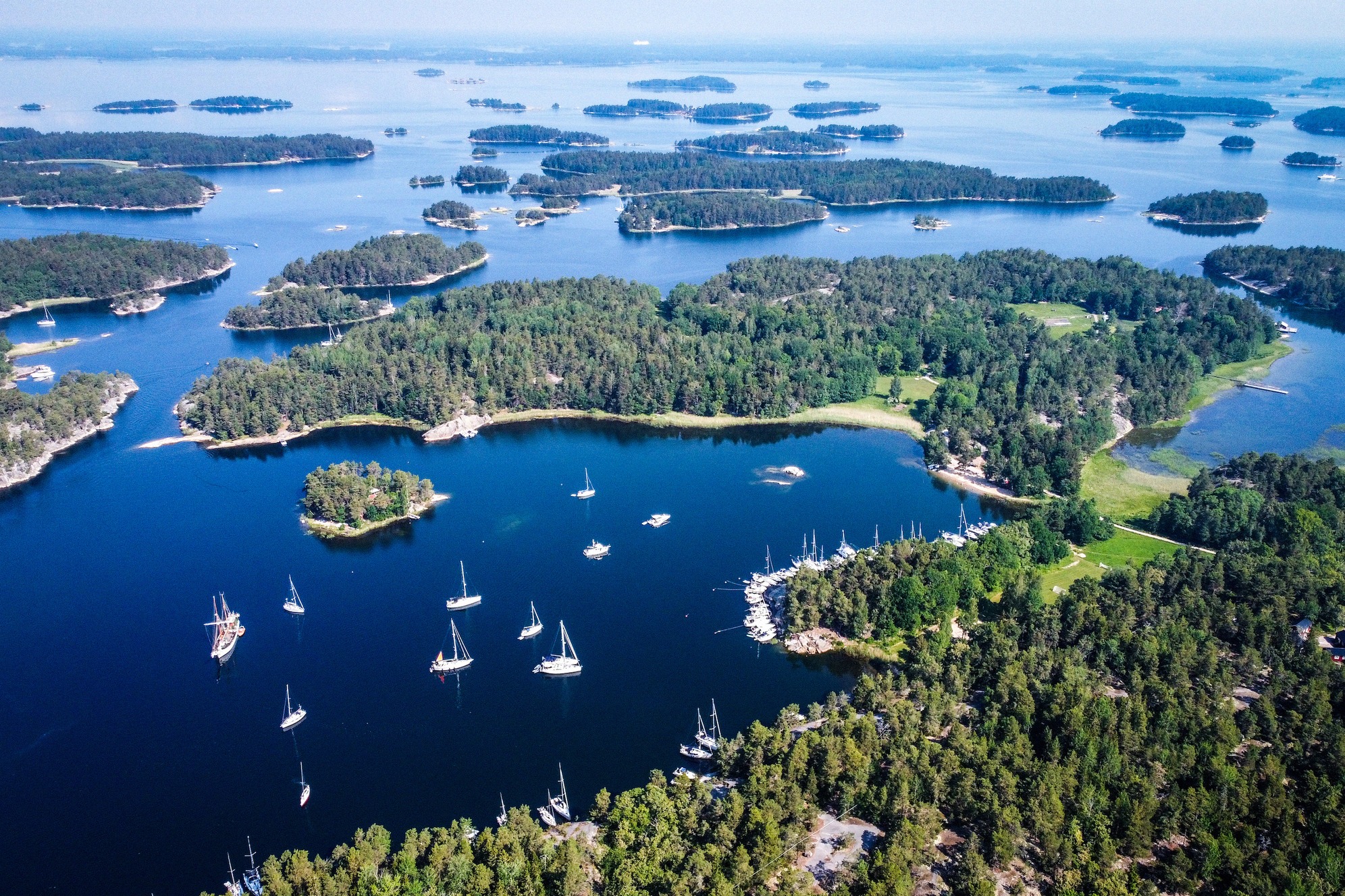 Tyypillinen ruotsalainen saaristomaisema punaisen puutalon kanssa rannalla. Katkennut puupaalu kohoaa matalasta vedestä, taustalla ruokoa ja puita. Idyllinen kesätunnelma.