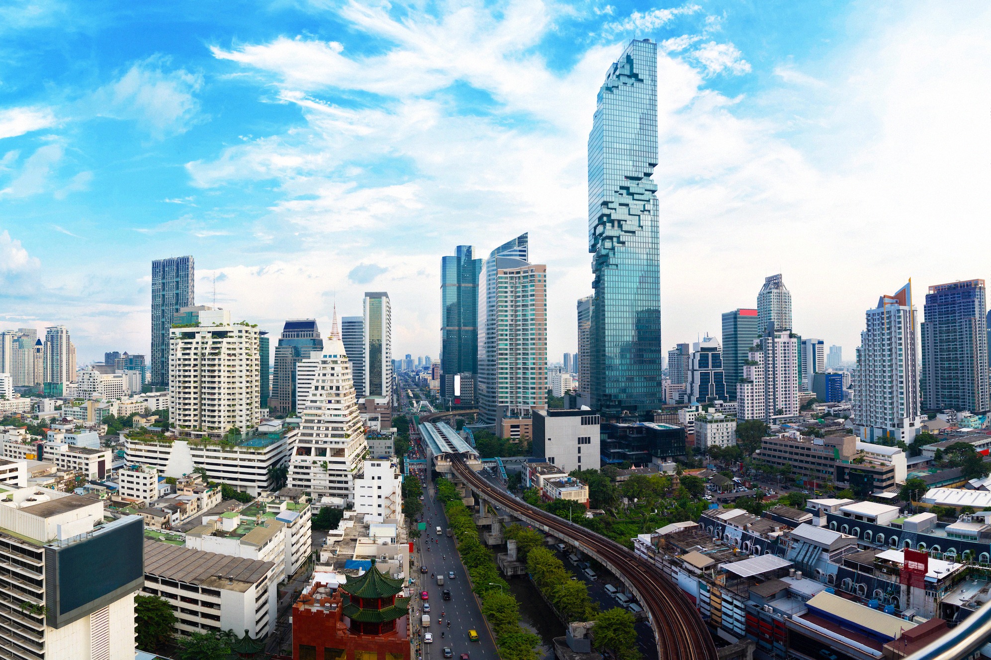 Vaikuttava panoraama Bangkokin silhuetista: näyttävä MahaNakhon Tower korkeimpana rakennuksena, Skytrainin silta etualalla ja moderneja pilvenpiirtäjiä sinisen taivaan alla.