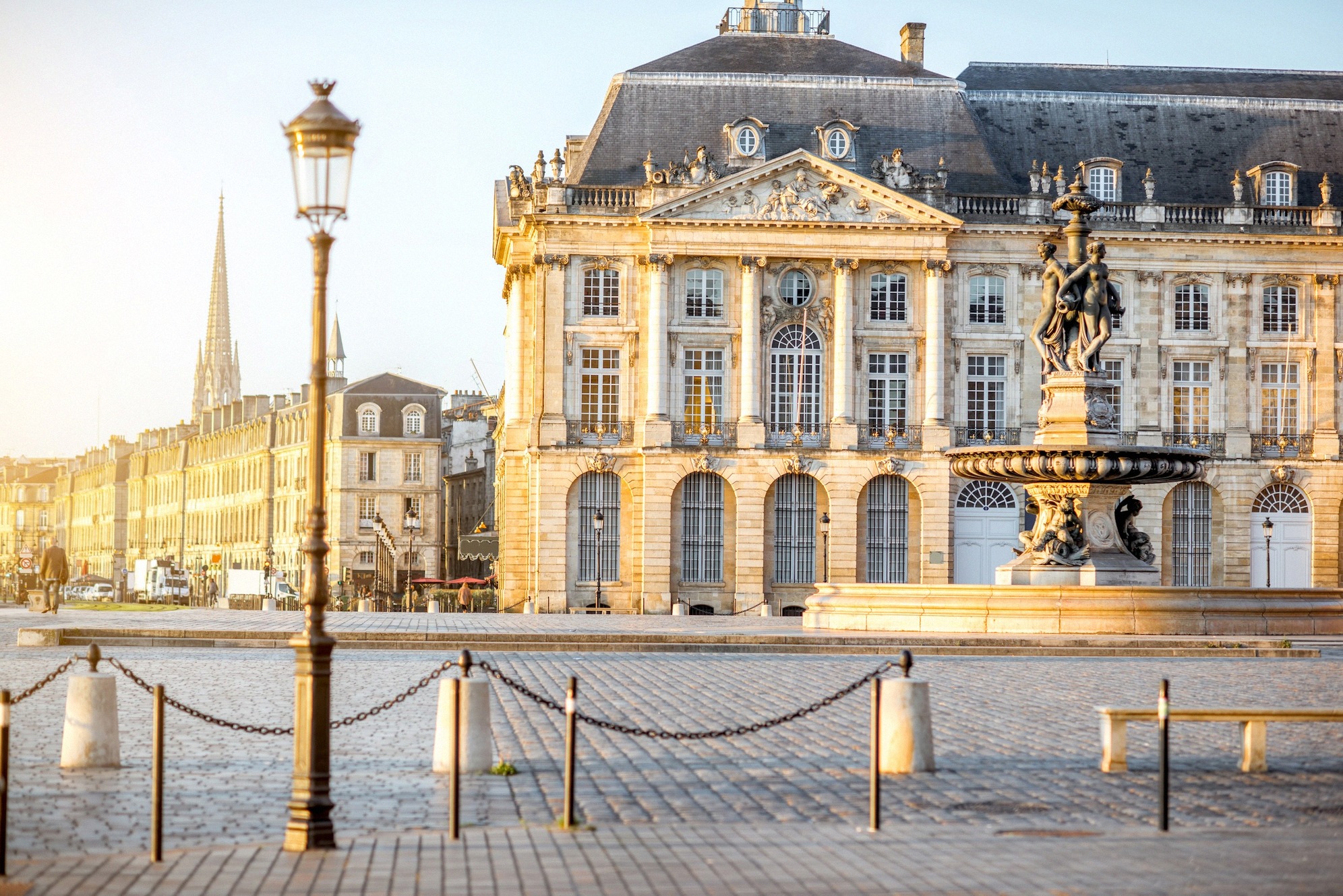 Place de la Bourse kultaisessa aamuvalossa: uusklassinen pörssirakennus ja kolmen sulottaren suihkulähde hehkuvat lämpimässä hiekkakivessä, taustalla Église Saint-Michelin torni - Bordeaux'n valokuvatuin näkymä.