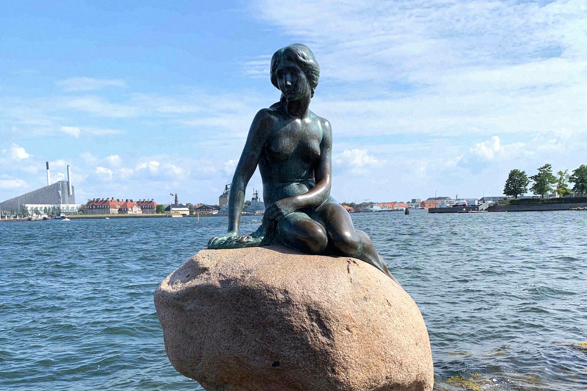 Maailmankuulu Pieni merenneito -pronssipatsas istuu kivellä Kööpenhaminan satamassa, taustalla kaupungin siluetti.