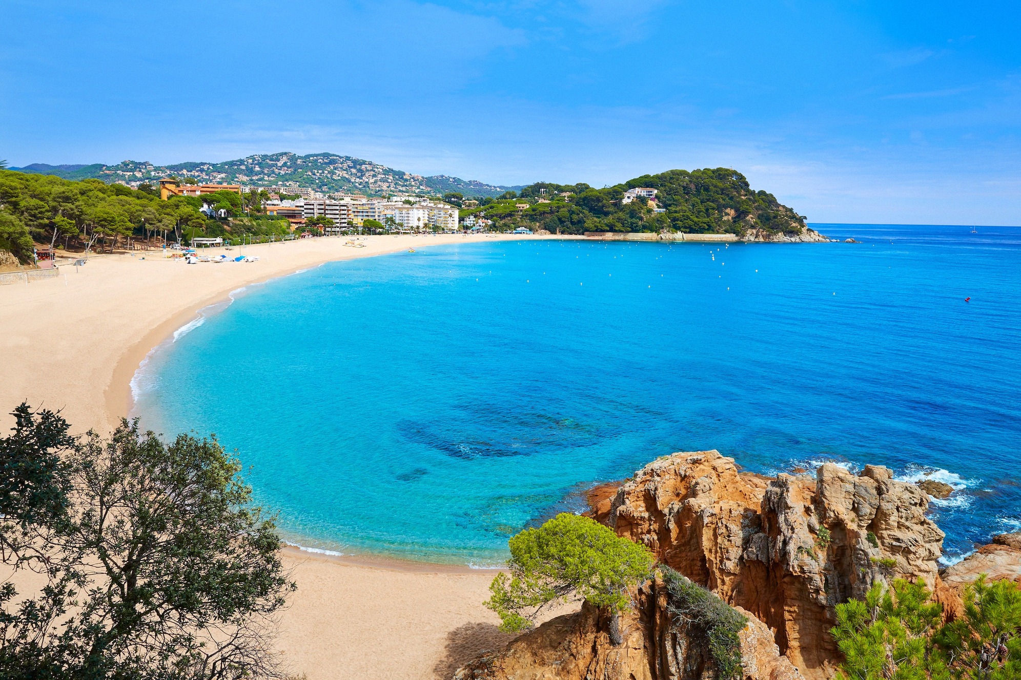 Platja Fenals Lloret de Marin luona Costa Bravalla: kaartuva hiekkaranta turkoosinsinisine vesineen, punaisine kalliomuodostelmineen ja metsäisine kukkuloineen taustalla.