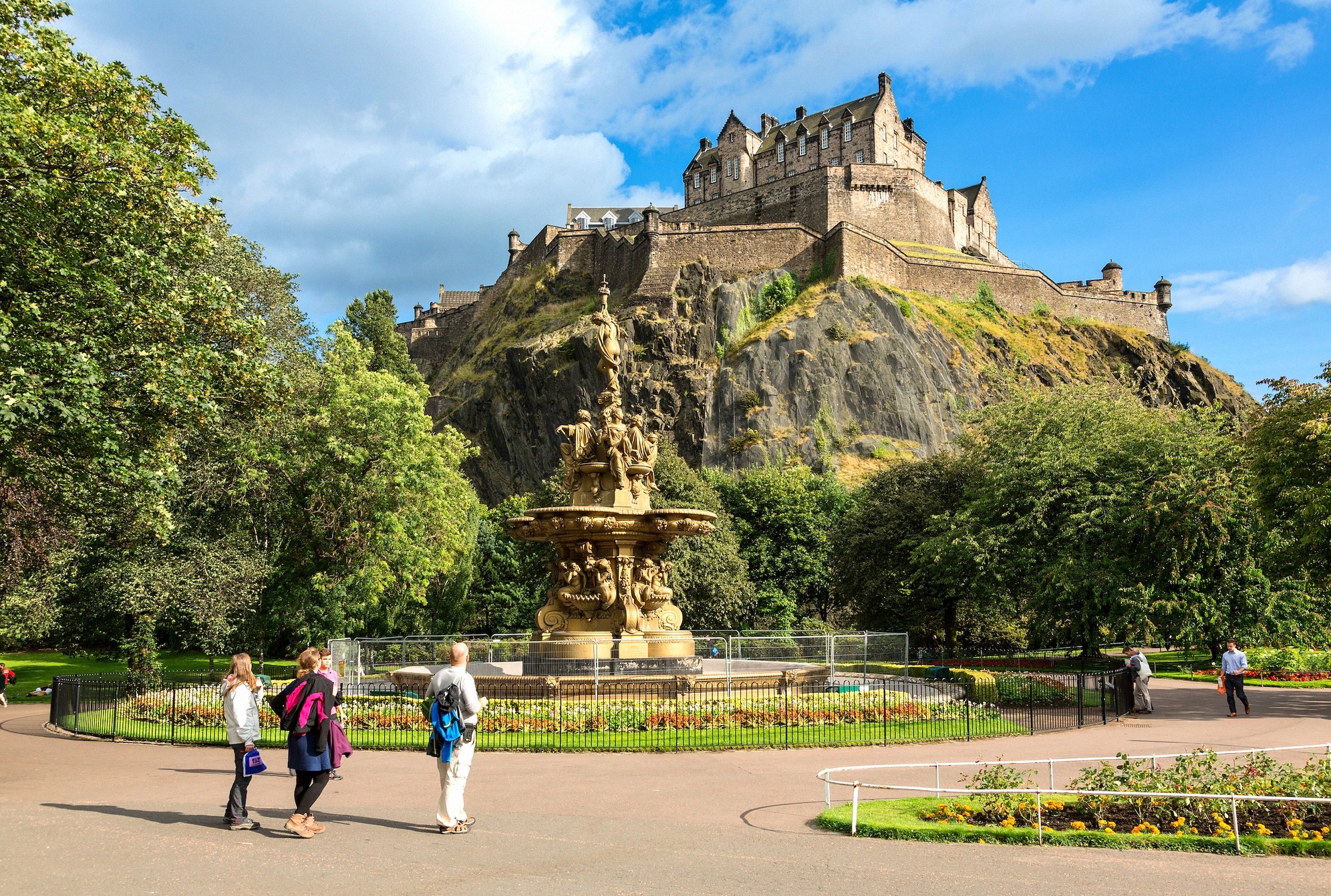 Edinburghin linna kohoaa linnakalliolla Princes Street Gardensin yllä; etualalla koristeellinen Ross-suihkulähde kesäisten kukkapenkkien ympäröimänä.