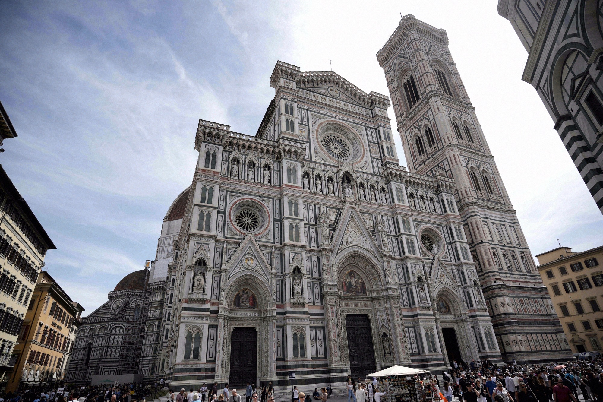 Firenzen tuomiokirkon valkovihreä marmorijulkisivu suoraan edestä kuvattuna, oikealla Giotton kellotorni ja turisteja aukiolla.