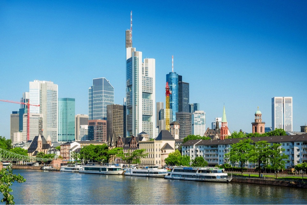 Frankfurtin klassinen pilvenpiirtäjäsilhuetti Commerzbank Towerineen heijastuu Mainiin, rannassa risteilyaluksia, taivas sininen.