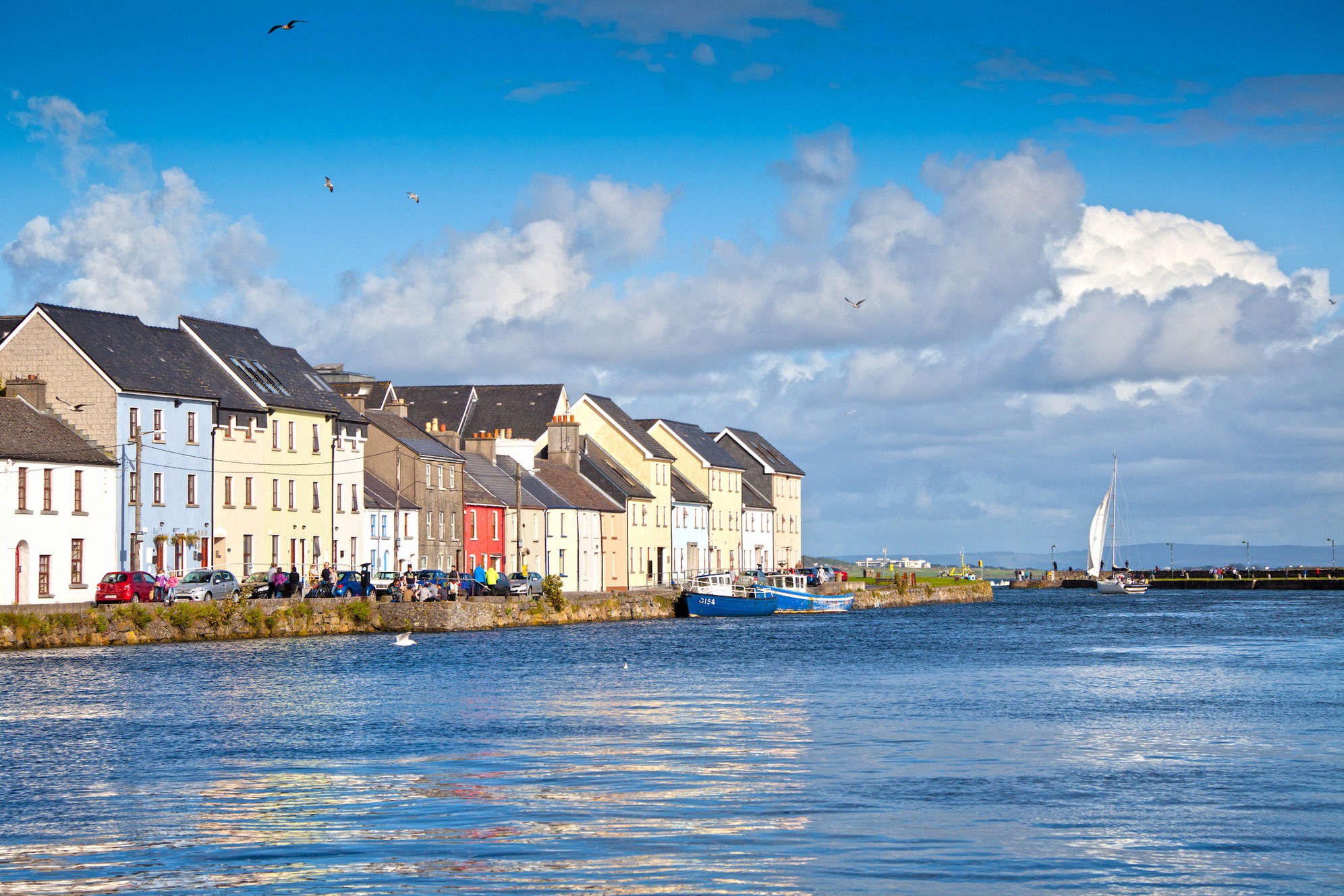 Galwayn Claddaghin kaupunginosa veden äärellä, rivi vaaleita julkisivuja, kalastusveneitä ja purjelaiva tyynellä Long Walkilla.