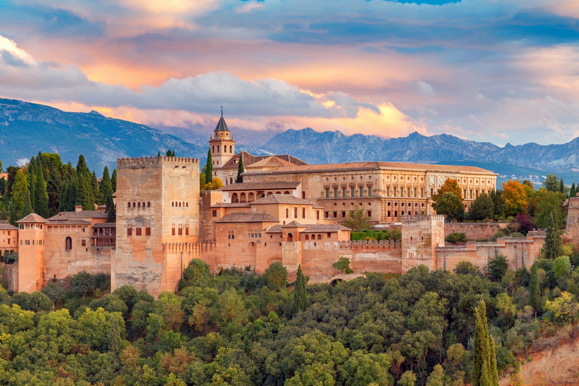 Granadan Alhambran linnoitus- ja palatsikokonaisuus lämpimässä iltavalossa: torneja, maurilaisia muureja ja Palacio de Carlos V lumihuippuisen Sierra Nevadan edessä.
