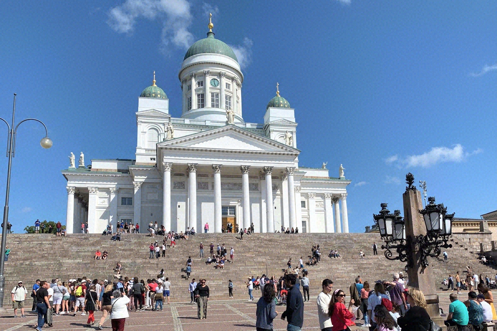 Helsingin Tuomiokirkko edestä kuvattuna Senaatintorilla kirkkaassa auringonpaisteessa. Leveillä portailla on runsaasti kävijöitä; vihreät kupolit ja uusklassinen pääjulkisivu hallitsevat näkymää.