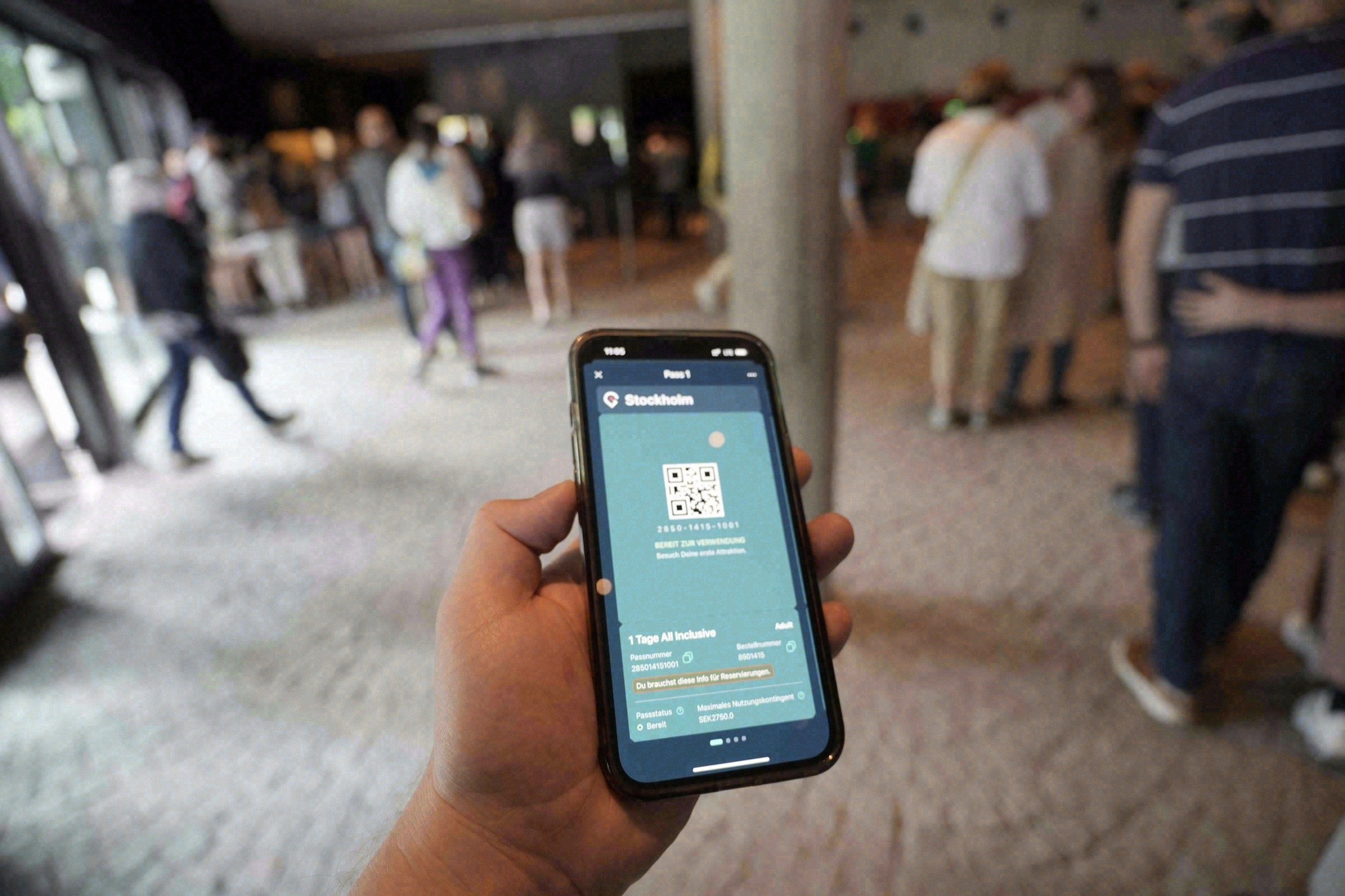 Kädessä älypuhelin, jonka näytöllä on digitaalinen city pass QR-koodeineen; taustalla vilkas museoaula.