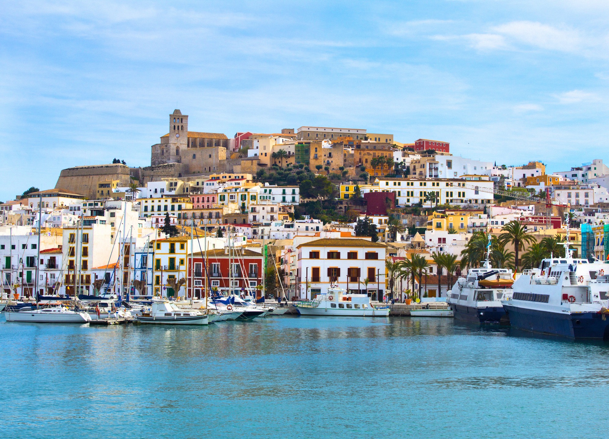 Näkymä satamasta värikkääseen alakaupunkiin ja mahtavaan Dalt Vila -linnoitukseen Ibizalla, purjeveneitä ja lauttoja laitureilla.