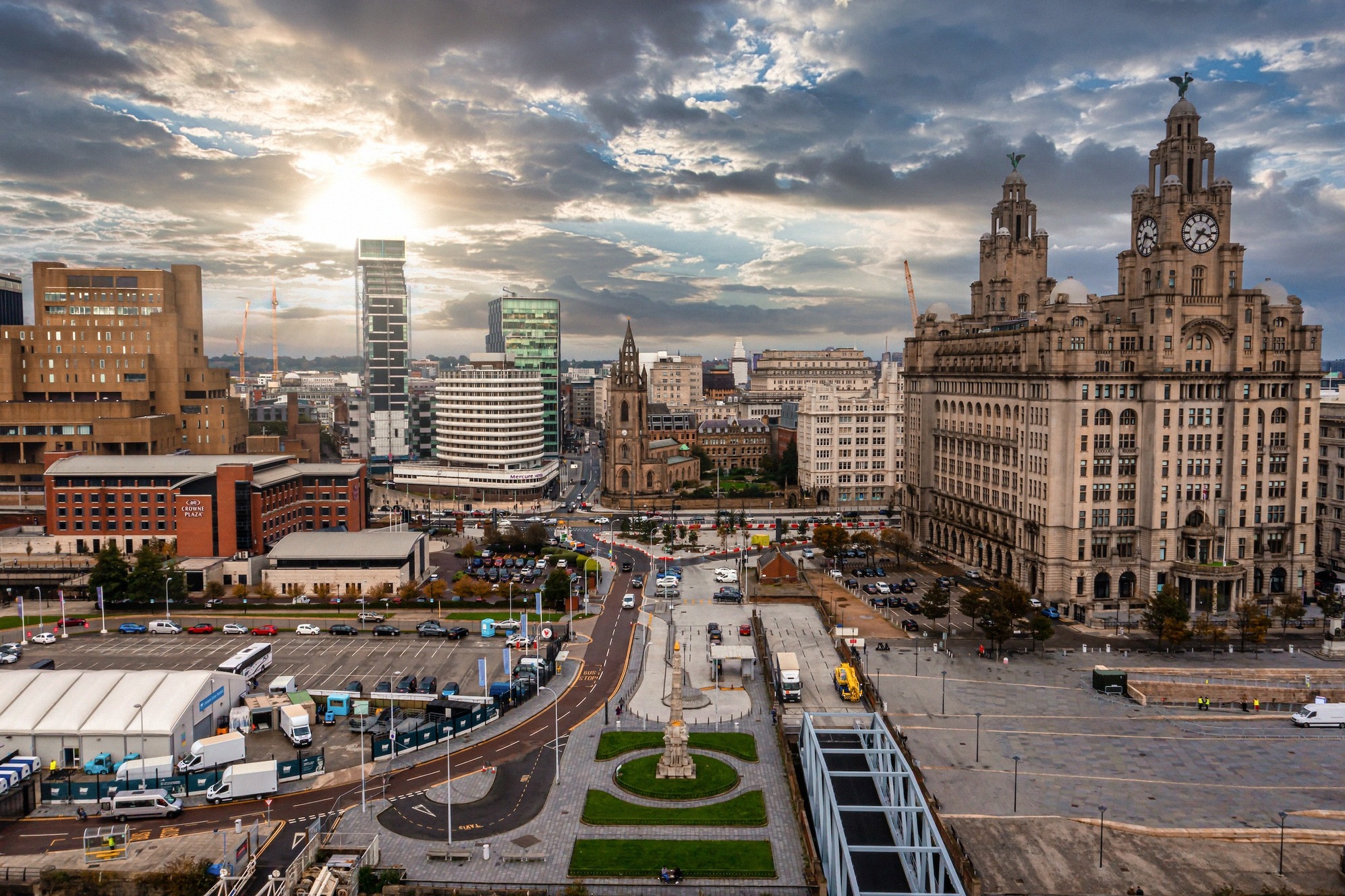 Ilmakuva Liverpoolin silhuetista, jossa ikoninen Royal Liver Building Pier Headilla, dramaattinen pilvitaivas laskevassa auringossa ja laaja kaupunkirakenne.