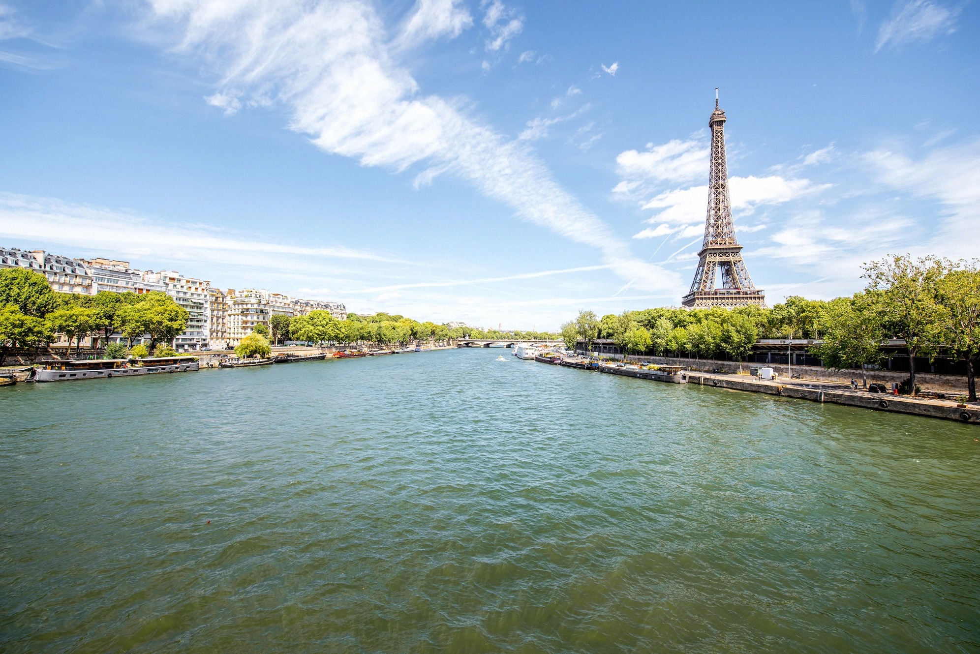 Laajakuva Seinestä Pariisissa, oikealla taustalla Eiffel-torni, rannoilla vihreitä puita ja asuntolaivoja kirkkaana kesäpäivänä.