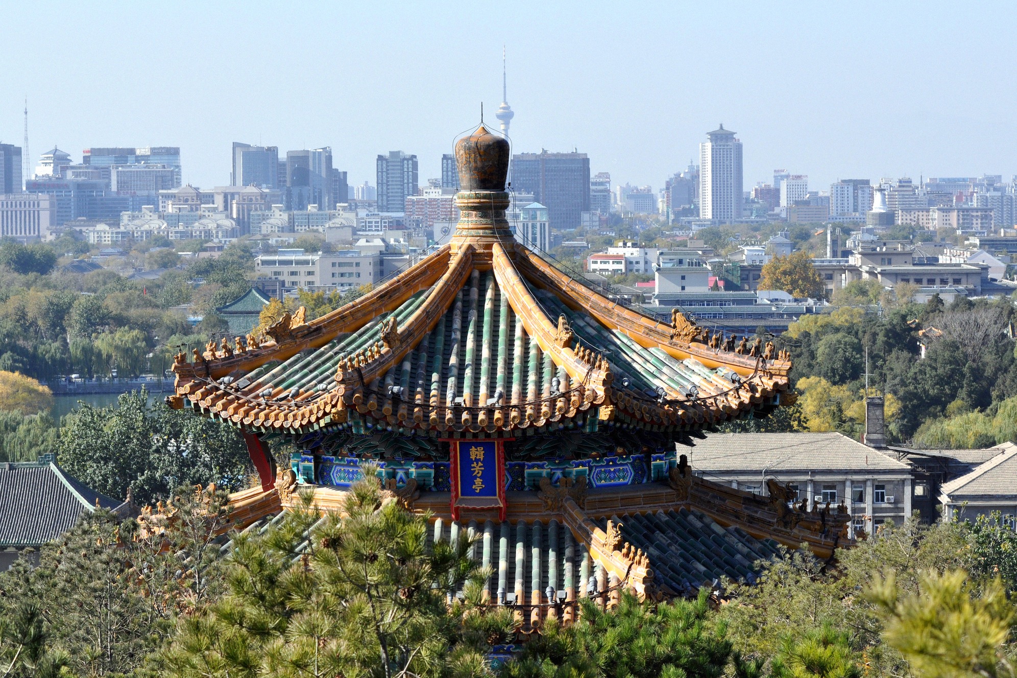 Lähikuva keisarillisesta paviljongista monivärisine tiilikattoineen Jingshanin puistossa, taustalla Pekingin kaupunkisilhuetti.