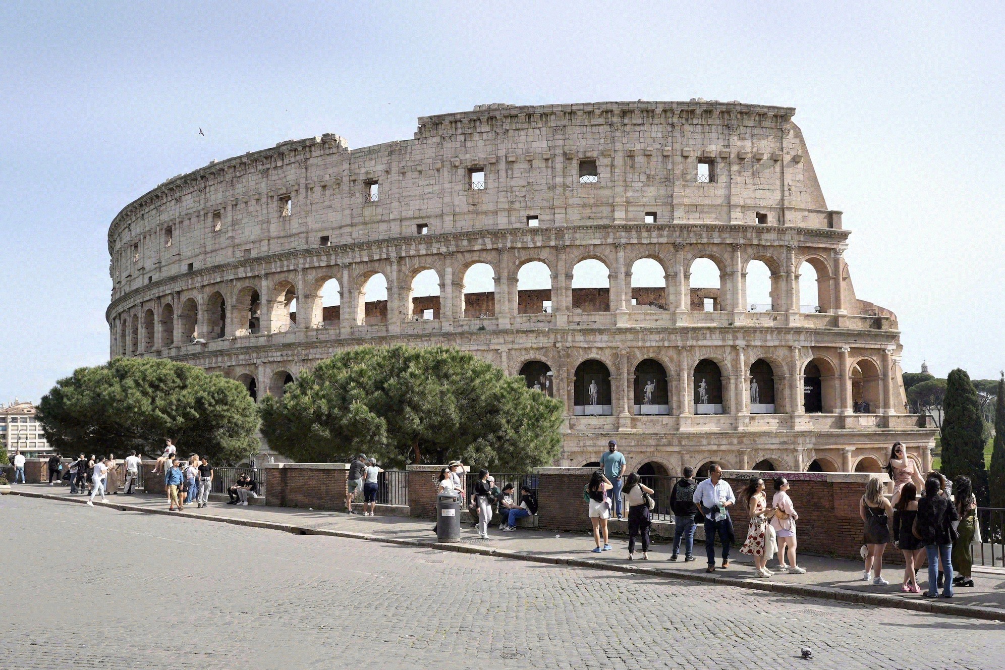 Näkymä Forum Romanumin yli antiikin pylväsriveineen; taustalla Colosseum. Runsaasti kävijöitä kivetyllä polulla kaivausalueiden reunalla, kesäinen tunnelma.