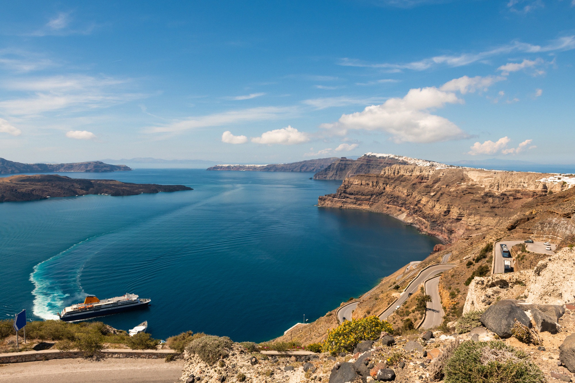 Laajakulmapanoraama Santorinin kalderasta, jossa lautta kulkee syvänsinisellä kraatterimerellä. Kaartuva serpentiinitie jyrkän rannikon vieressä, Nea Kamenin ja Thirasian saaret sekä valkoinen Oia taustalla täydentävät dramaattisen tulivuorimaiseman.