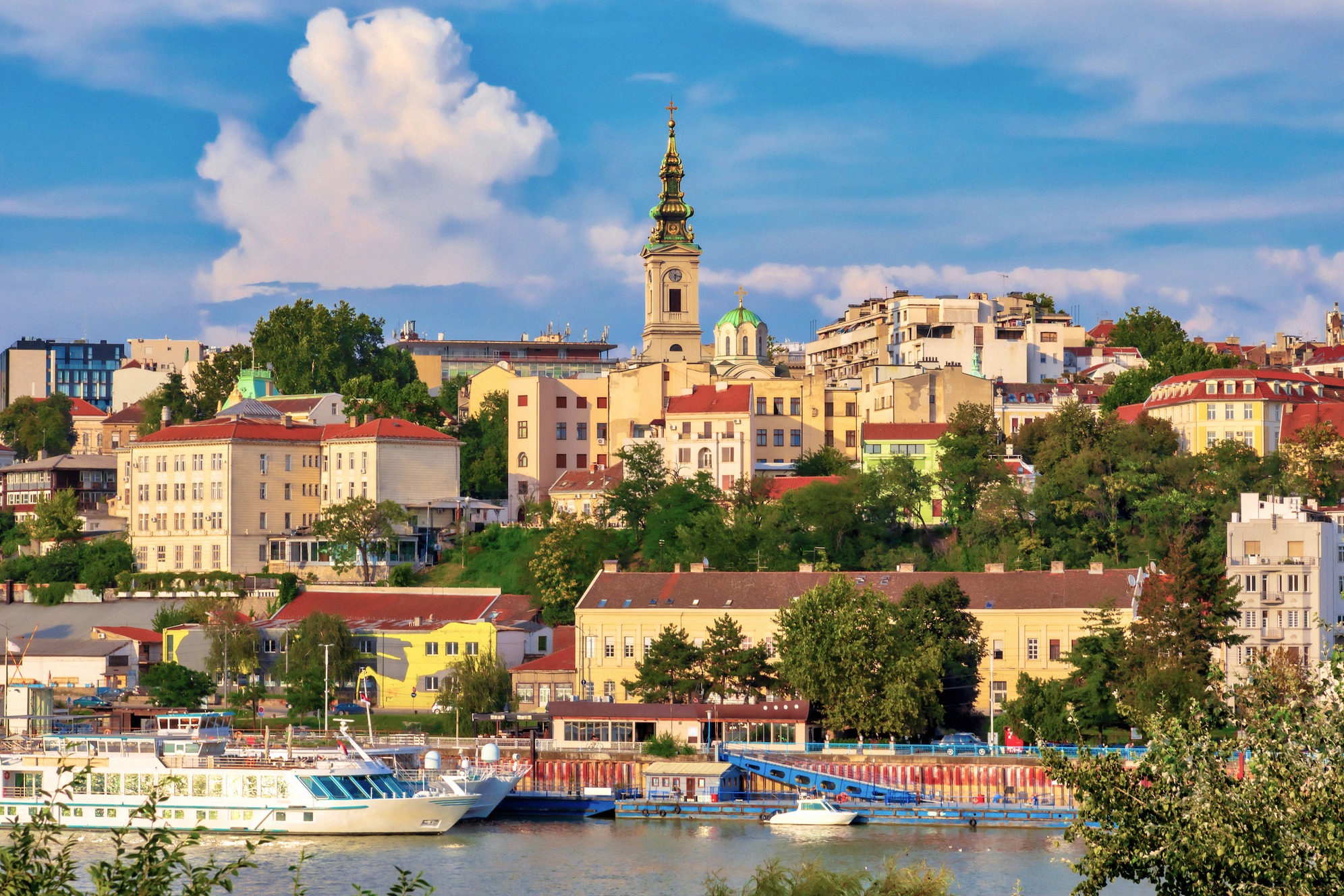 Belgrad Savajoelta katsottuna, vanhankaupungin värikkäät julkisivut ja vihreät kukkulat kirkontorneineen, jokivenheitä rannassa sinisen kesätaivaan alla