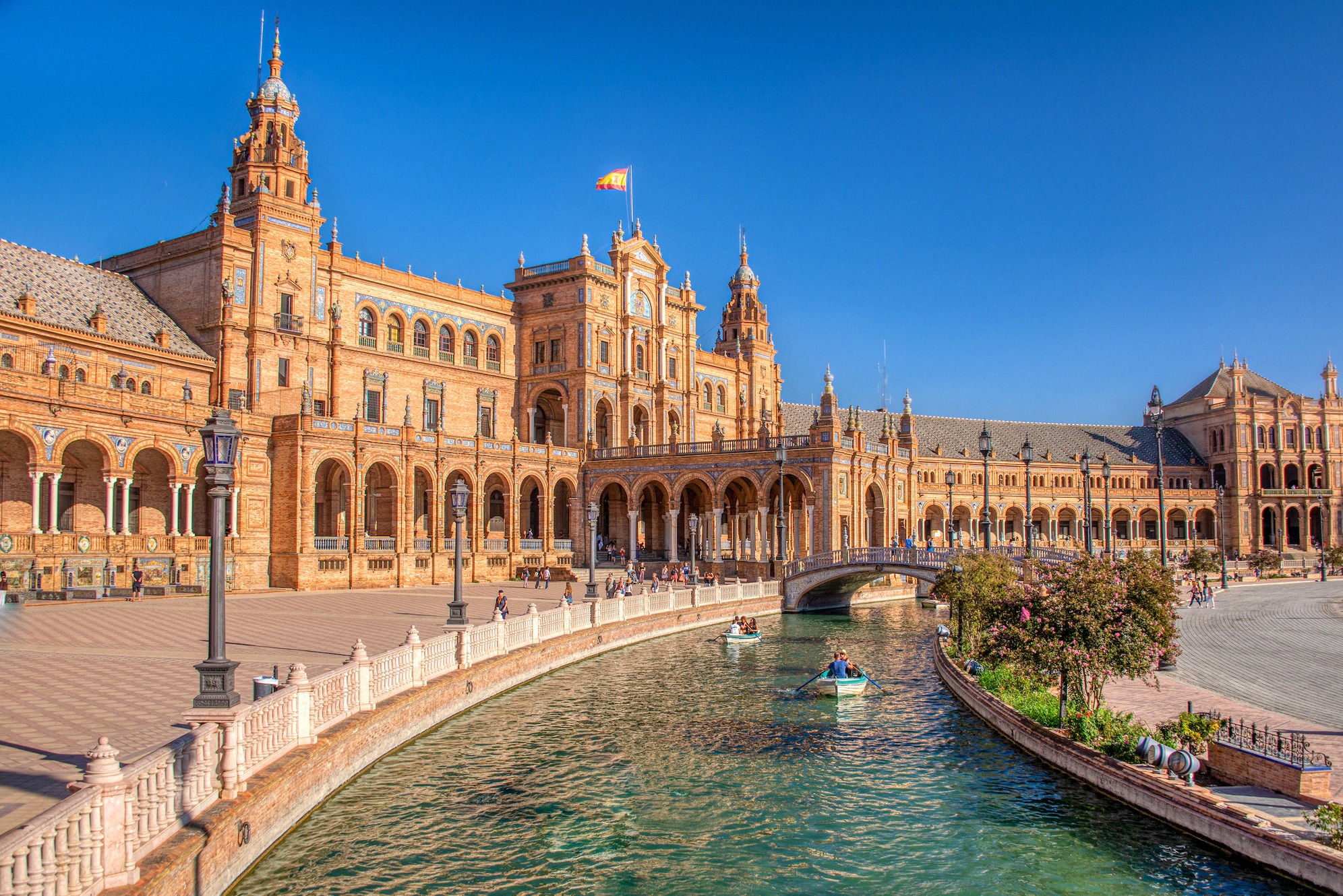 Sevillan upea Plaza de España puolikaaren muotoisine, uusmudéjar-tyylisine palatsirakennuksineen. Edessä olevalla kanavalla kulkee pieniä soutuveneitä ja Espanjan lippu liehuu keskusrakennuksen yllä, ikoninen kuva Iberian etelästä.