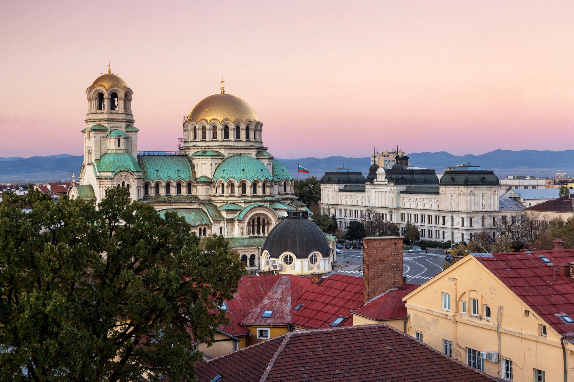 Aleksanteri Nevskin katedraalin kultainen kupoli hehkuu iltaruskossa Bulgarian pääkaupungin Sofian kattojen yllä, taustalla kansalliskokouksen rakennus.
