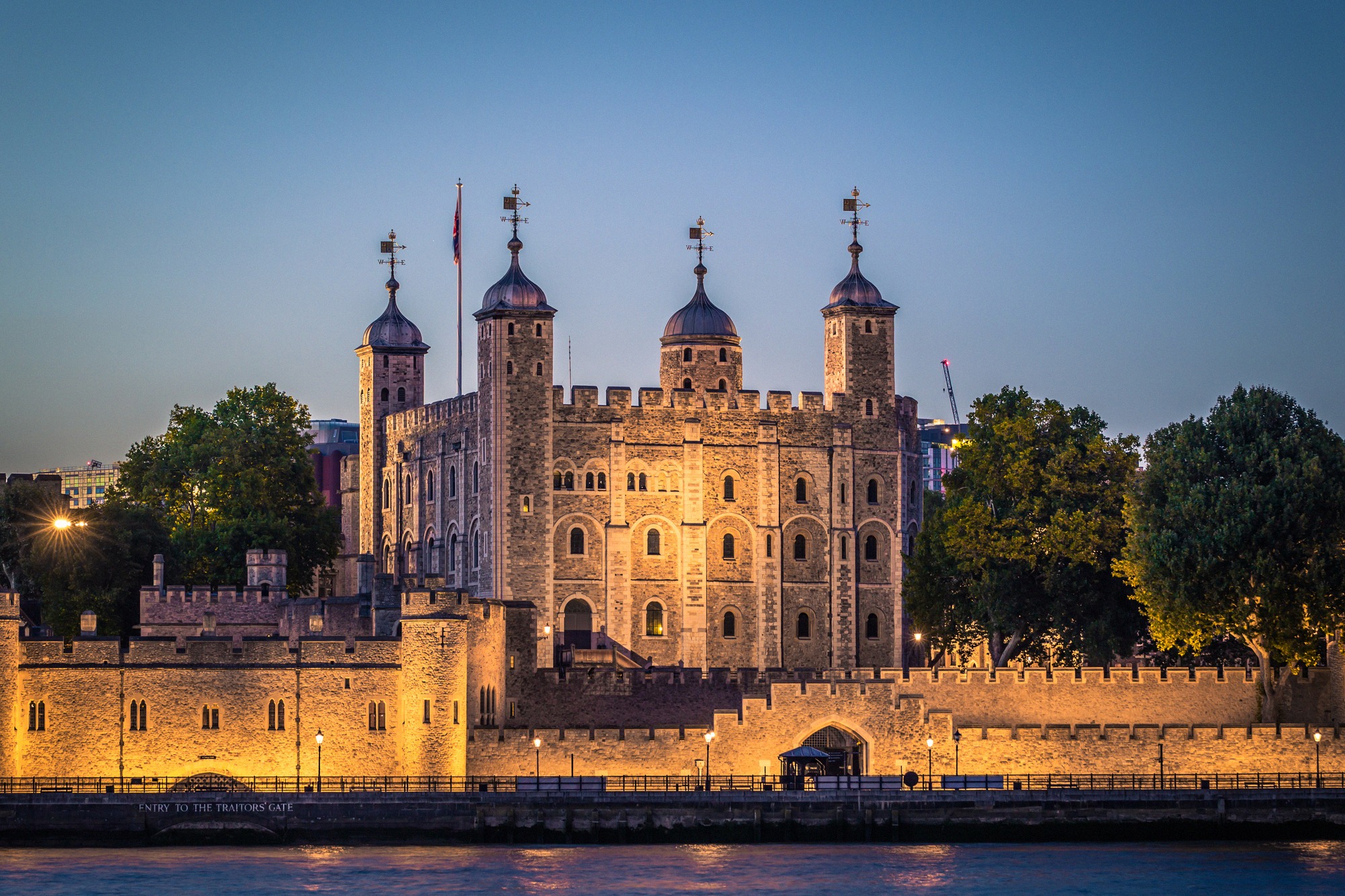 Tower of London Thamesin varrella sinisessä iltavalossa, valaistu White Tower kohoaa puolustusmuurin yllä.